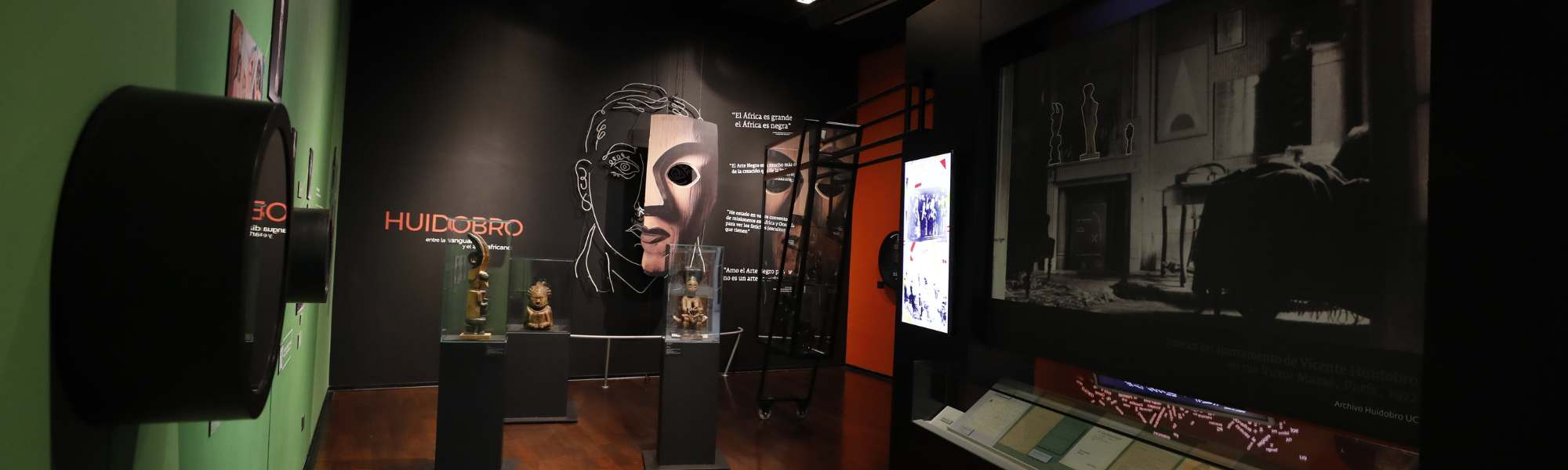 Vista general de sala de exposiciones con dibujo en neón del rostro de Vicente Huidobro al fondo y distintos objetos y gráficas